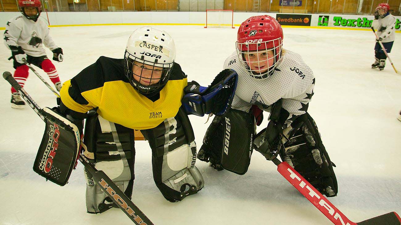 Två barn som står i ishallen med hockeyutrustning på sig.